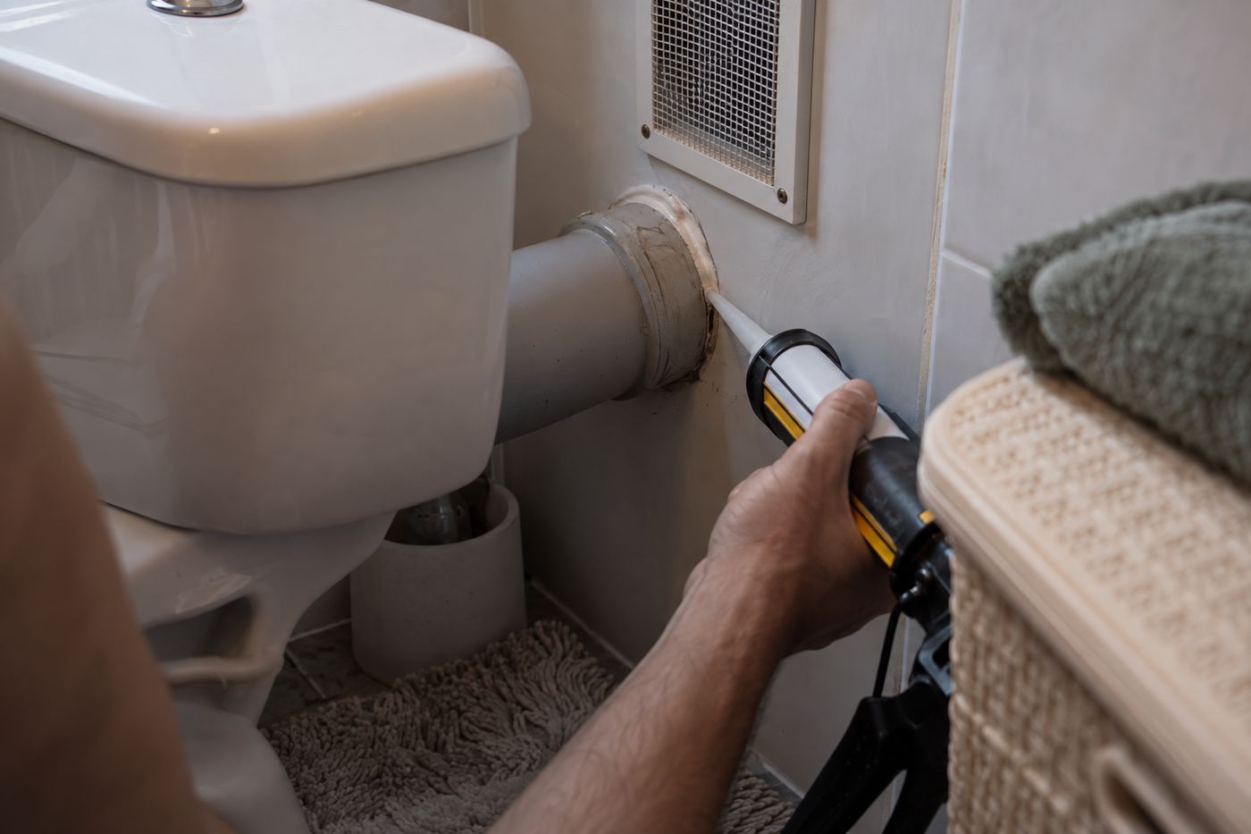 Silicone caulk sealing a gap around a pipe behind a toilet, with mesh over a vent