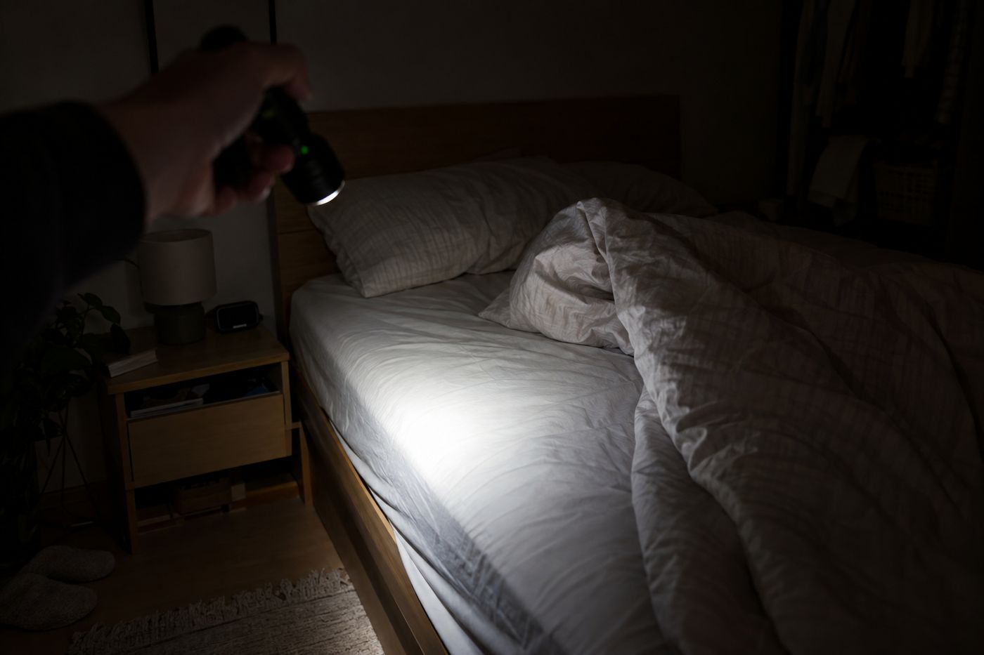 Checking a bed at night with a flashlight: blanket pulled back over a light-colored sheet, alarm clock on the nightstand